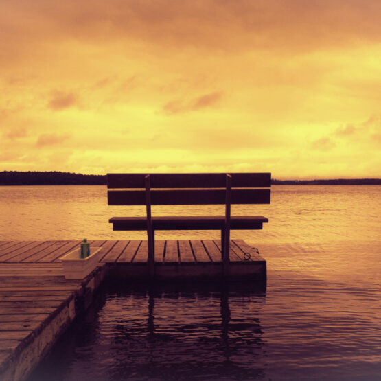 A serene lake scene at sunset with a wooden dock bench overlooking calm waters under a warm, golden sky.