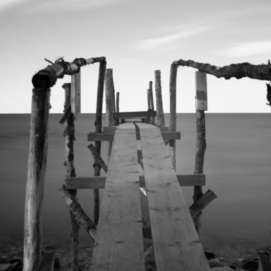 A black and white infra-red photograph showing a rustic, makeshift wooden dock leading out into a calm body of water, framed by wooden posts on either side, under a hazy sky.