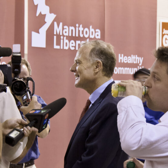 Manitoba Liberal Party leader Dr. Jon Gerrard smiling while being interviewed by multiple journalists with cameras and microphones, with banners reading 'Manitoba Liberal' and 'Jon Gerrard' in the background, and another man, Press Secretary David Shorr, in the foreground drinking from a bottle.
