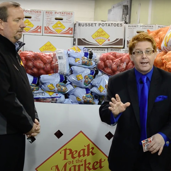 Peak of the Market's Larry McIntosh and Illusionist Brian Glow in front of a display of stacked bags of potatoes and onions, one in a black jacket, standing with his hands clasped, looking at the other man in a blue suit who is explaining something with an expressive gesture, in a warehouse setting with a 'Peak of the Market' sign.
