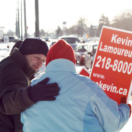 A smiling man, candidate Kevin Lamoureux in a winter hat and coat, embracing another person in a blue jacket from behind, both standing on a snowy roadside next to a red election sign for Kevin Lamoureux.