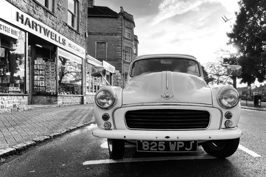 Monochrome image of a classic white Morris Minor parked on a cobblestone street in front of Hartwells, a bicycle hire shop. The setting has an old-world charm, with vintage architecture and a plane flying in the distance under a sky with soft clouds.