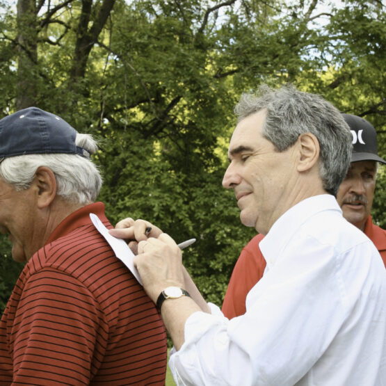 Michael Ignatieff, seen from the back on the left, is having his shirt signed by a man in a white shirt; both are at an outdoor event with trees in the background.